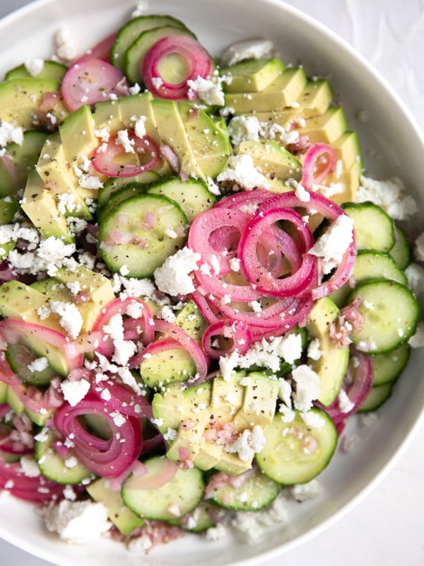A bowl of cucumber onion salad in a large shallow white bowl with red onions, creamy avocado, thinly sliced cucumber, crumbled feta cheese, and light red wine vinaigrette.