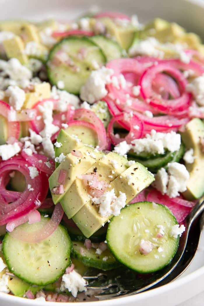 Close up image of Large white salad plate filled with sliced cucumbers, avocado, and pickled onions covered in crumbled casero cheese and homemade vinaigrette.