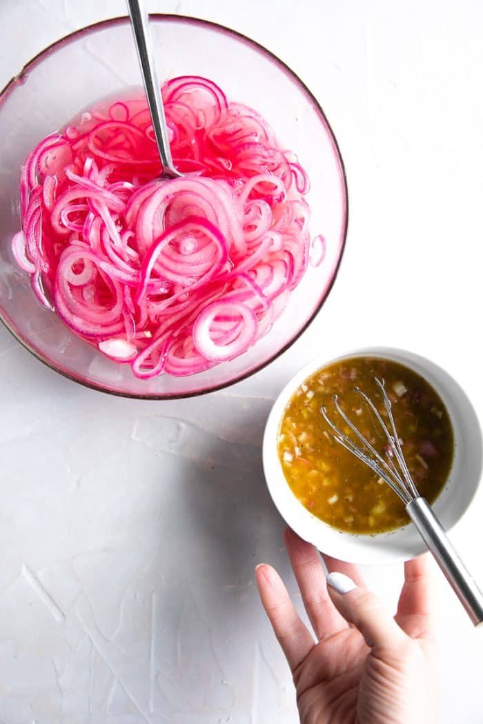 Two bowls on a white surface. The first bowl is filled with pickled red onions and the second bowl is filled with a red wine vinaigrette.