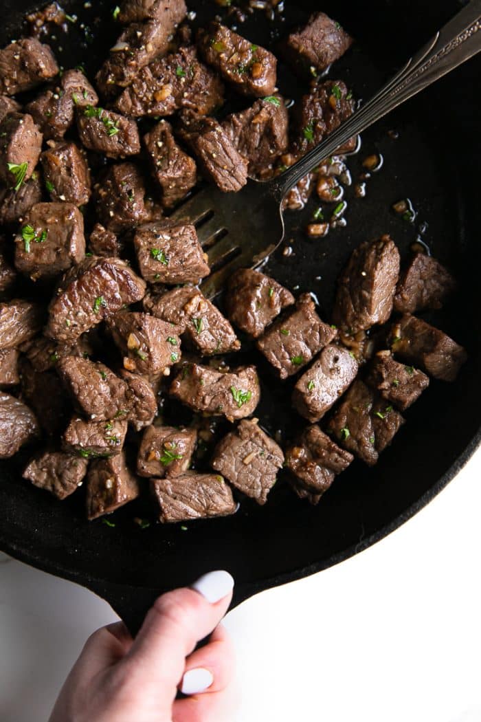 Person holding the handle of a cast iron skillet filled with garlic butter sirloin steak bites.