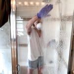 A woman standing in front of a glass shower door spreading chemicals for cleaning hard water build up