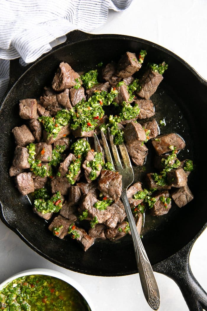 Overhead image of a large black cast iron skillet filled with cooked steak bites drizzled with chimichurri sauce.
