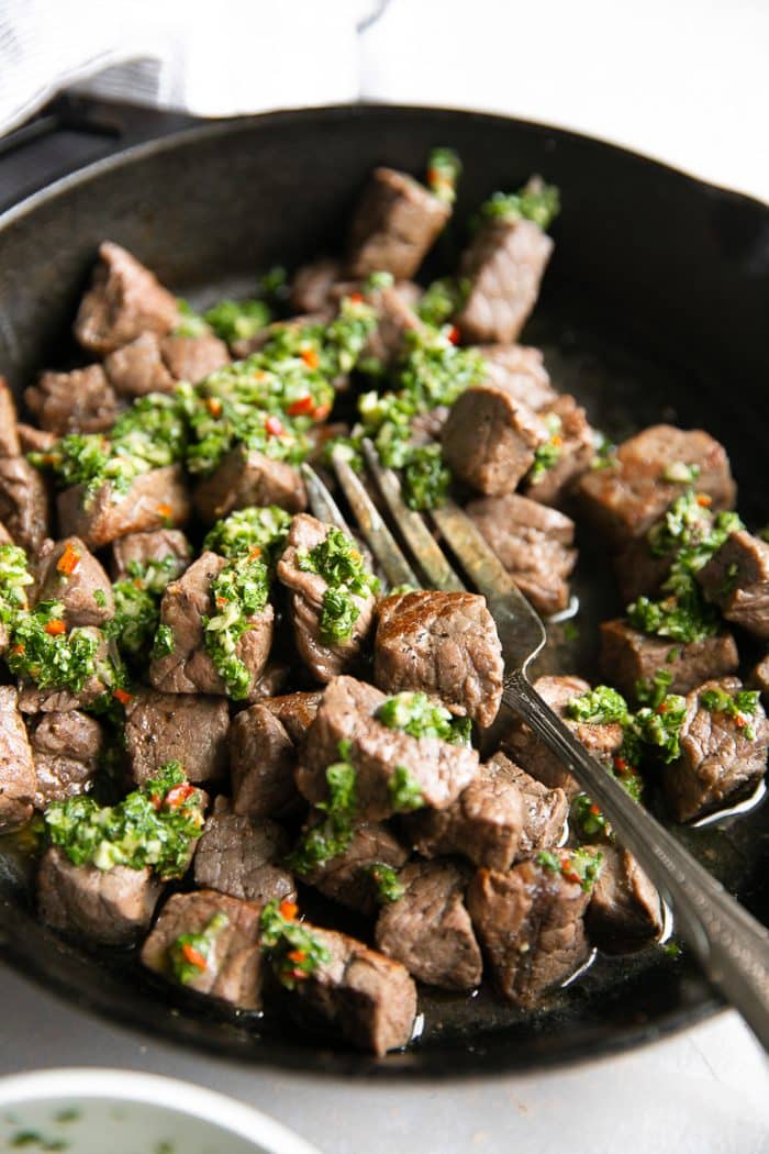 Large silver fork scooping up steak bites covered with chimichurri sauce from a black cast iron skillet.