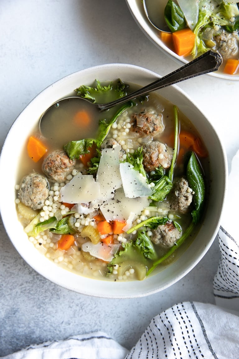 White bowl filled with clear chicken broth, small pasta, mini meatballs, carrots, and fresh greens.