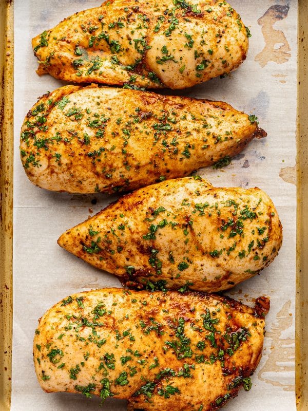 Four golden-brown oven baked chicken breasts sprinkled with finely chopped fresh parsley and arranged in a row on a parchment-lined gold baking sheet.