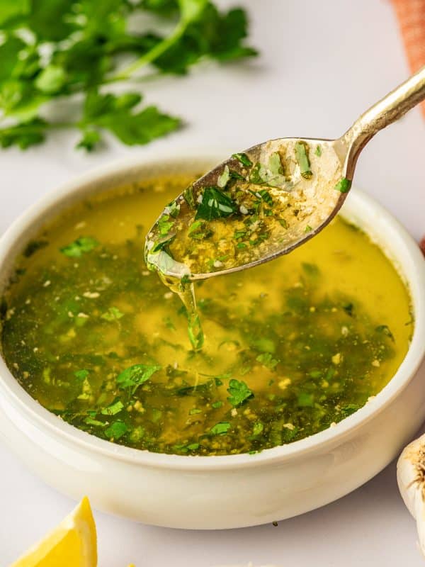 A spoon is lifted above a white ceramic bowl of garlic butter sauce, with sauce dripping from the spoon back into the bowl; chopped parsley is scattered on the surface of the sauce, lemon wedges and garlic bulbs sit in the foreground, and an orange fringed napkin and parsley leaves are visible in the background.