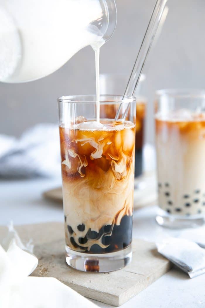 Pouring milk into a tall glass filled with cooked tapioca pearls, strong black tea, and ice.