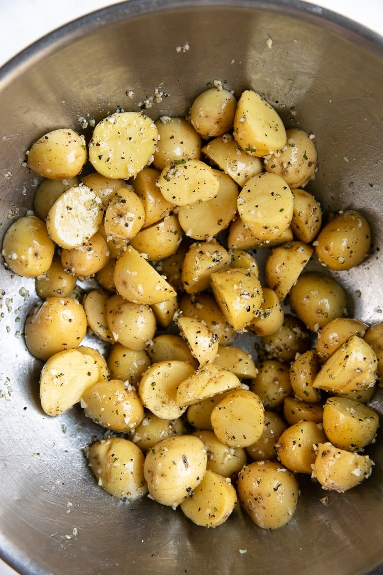 Large mixing bowl filled with baby potatoes covered in olive oil, garlic, salt, pepper, and minced rosemary.