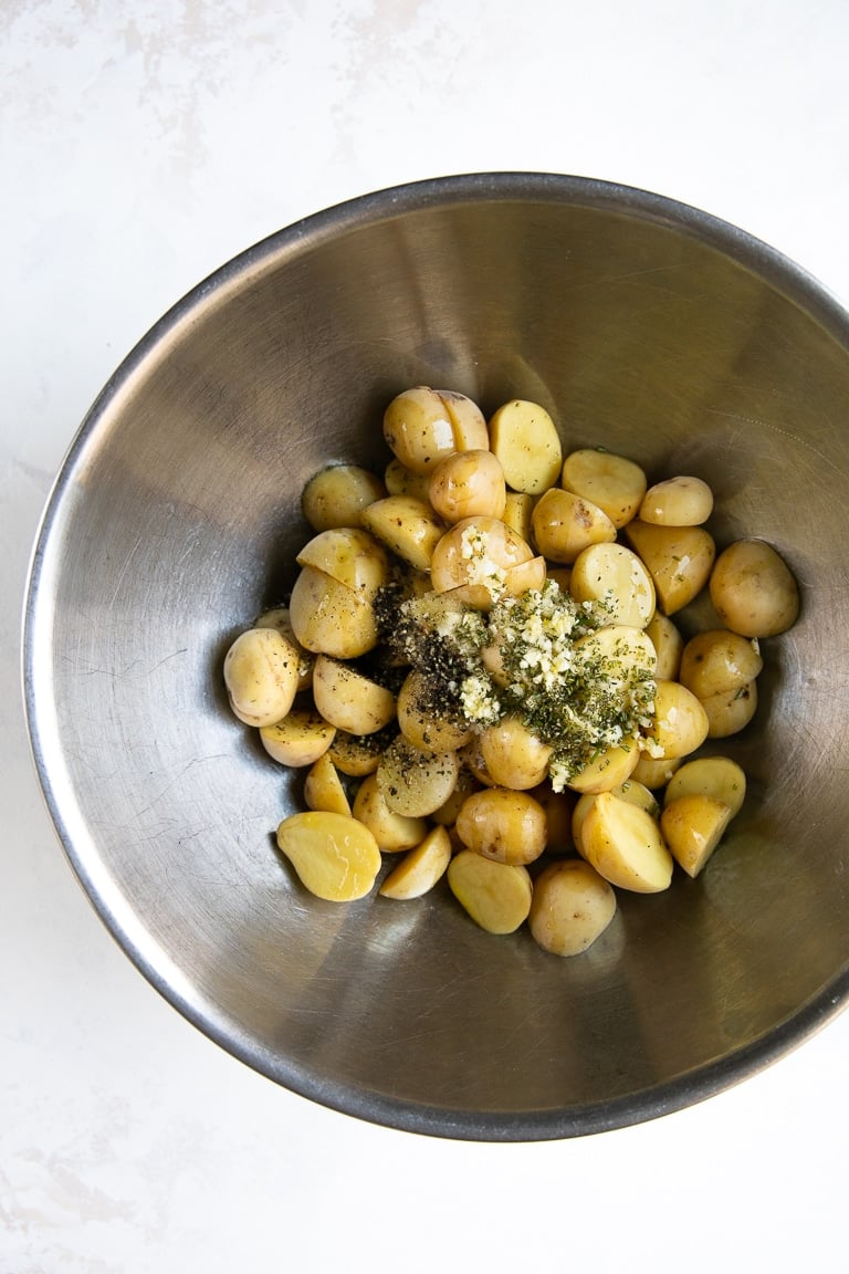 Large mixing bowl filled with baby potatoes, salt, pepper, olive oil, and garlic.