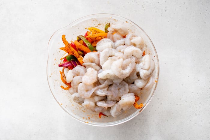 Raw peeled shrimp added to a bowl of seasoned peppers and onions.