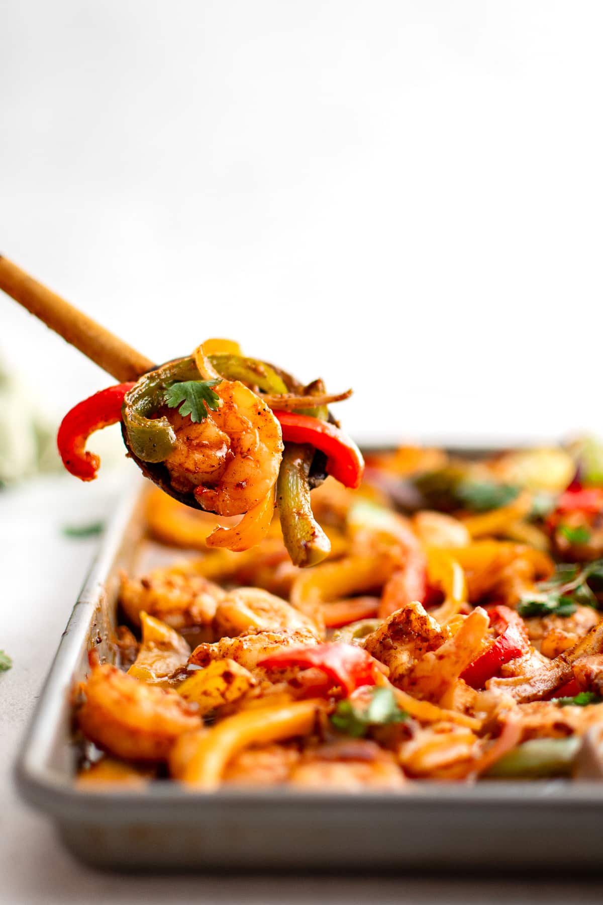 Wooden spoon lifting shrimp and bell peppers from a sheet pan of shrimp fajitas