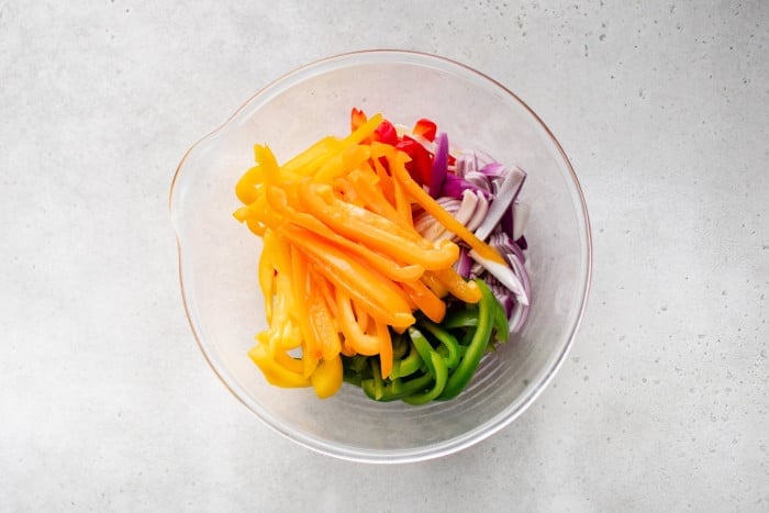 Sliced red, yellow, orange, and green bell peppers with red onions in a clear mixing bowl.