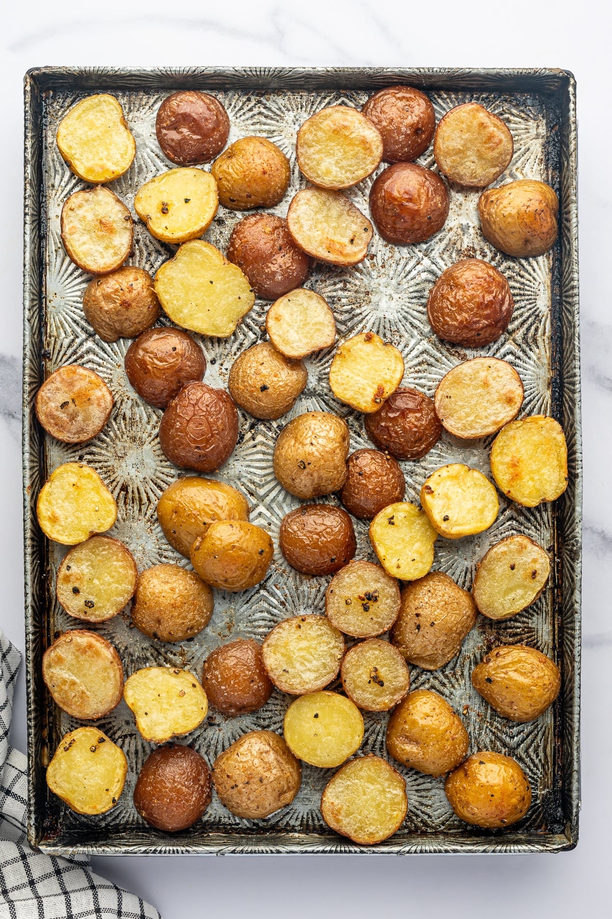 Overhead view of partially roasted red and Yukon Gold baby potatoes spread across a rectangular silver metal baking sheet with dark edges, with lightly wrinkled skins and cut surfaces beginning to turn golden brown. A black-and-white checkered towel is partially visible at the lower left.