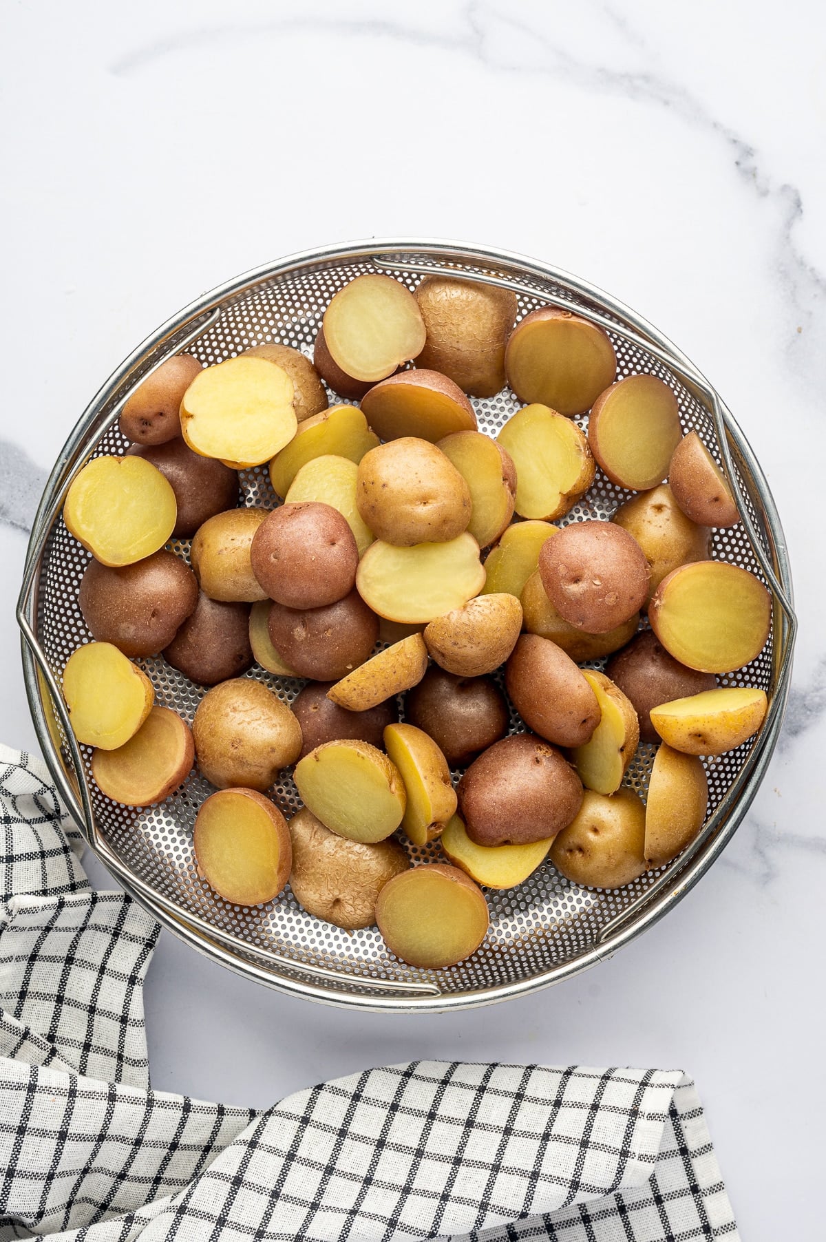Overhead view of parboiled red and Yukon Gold baby potatoes draining in a round silver metal colander on a white marble surface, with a black-and-white checkered kitchen towel arranged beneath the colander.
