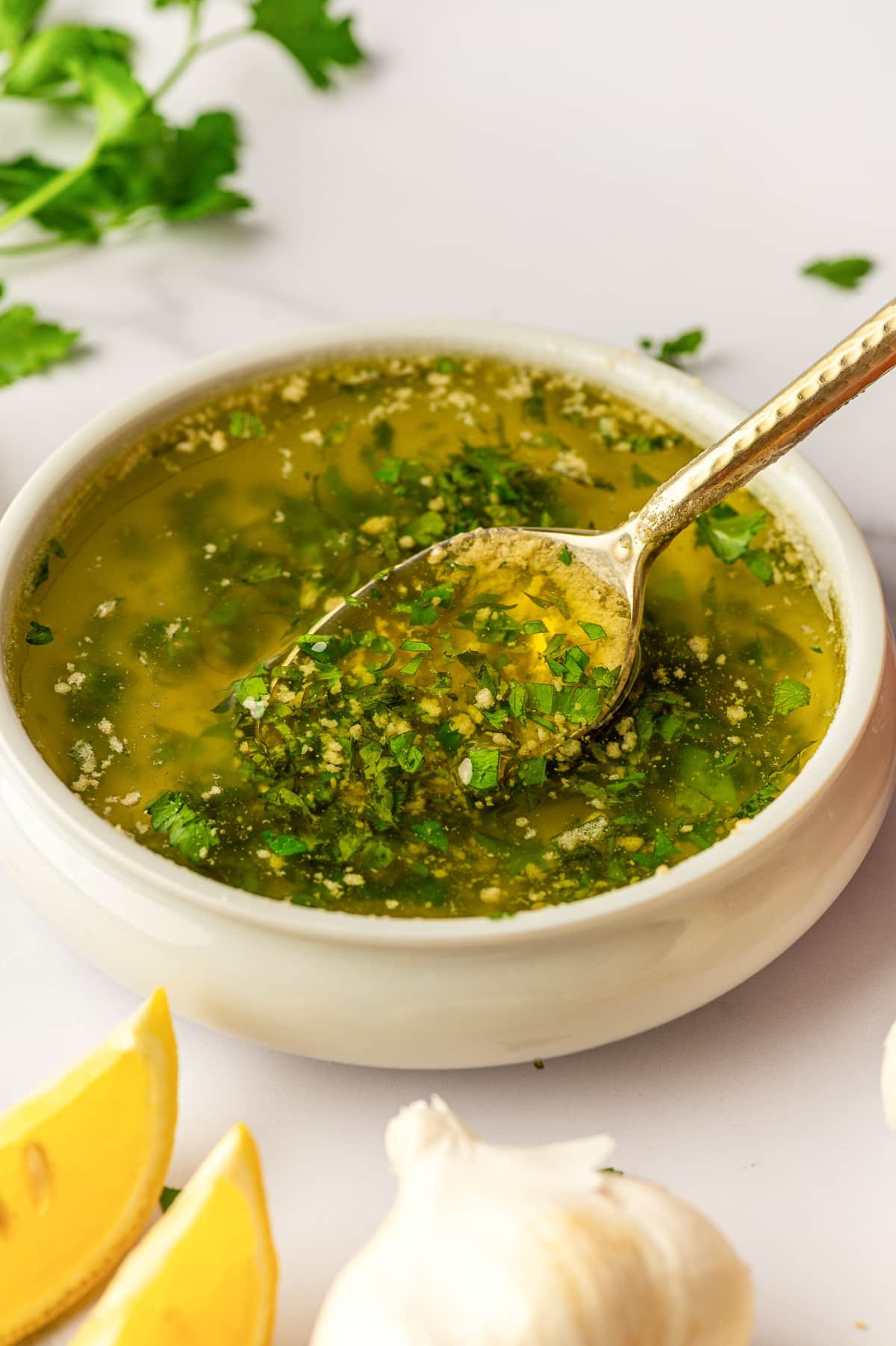 Close-up of a white ceramic bowl filled with golden garlic butter sauce with chopped parsley throughout, with a gold spoon resting across the bowl and holding sauce; blurred parsley leaves appear in the background, and lemon wedges and garlic bulbs sit near the bottom edge on a light countertop.