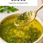 Vertical Pinterest pin with a large white text box at the top that reads “5-Minute GARLIC BUTTER SAUCE” in bold black letters and “vegetarian and gluten-free” in smaller black text underneath. Below the text box is a close-up photo of a white ceramic bowl filled with golden garlic butter sauce flecked with chopped parsley. A gold spoon is lifted above the bowl with sauce dripping back in. Lemon wedges and whole garlic bulbs sit near the bottom edge, a soft-focus parsley bunch is in the background, and an orange fringed napkin is visible at the right. Small black text at the bottom reads “the forked spoon.”
