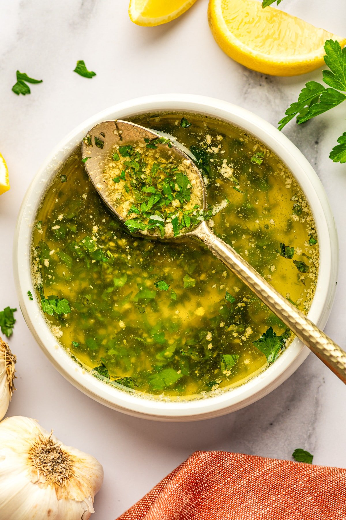 A white ceramic bowl filled with glossy garlic butter sauce flecked with chopped parsley sits on a light marble surface, with a silver spoon resting in the bowl and coated in sauce; lemon wedges and parsley sprigs surround the bowl, with whole garlic bulbs at the bottom edge and an orange fringed napkin in the lower right corner.