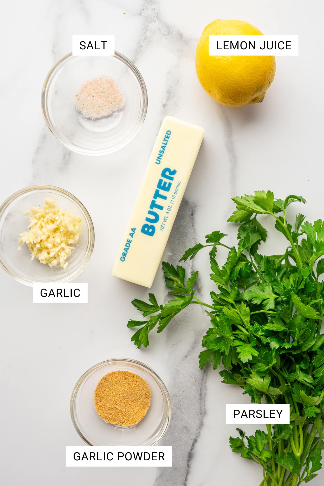 Overhead ingredients photo on a white marble background with white text labels in black font that read “SALT,” “LEMON JUICE,” “GARLIC,” “GARLIC POWDER,” and “PARSLEY”; a small glass bowl of pink salt sits under the SALT label, a whole lemon sits under the LEMON JUICE label, a small glass bowl of chopped garlic sits under the GARLIC label, a small glass bowl of garlic powder sits under the GARLIC POWDER label, and a large bunch of fresh parsley sits under the PARSLEY label, with a stick of butter in the center printed with blue “BUTTER” and “UNSALTED.”