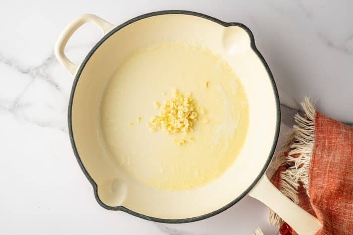 Overhead view of a cream colored enamel saucepan with a black rim on a white marble countertop, filled with melted butter and a small mound of minced garlic in the center; an orange fringed linen napkin sits to the right of the pan.