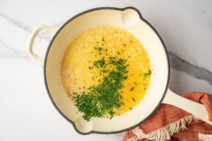 Overhead view of a cream colored enamel saucepan filled with a golden melted butter mixture speckled with seasoning, topped with a pile of chopped fresh parsley in the center; the pan sits on a white marble countertop with an orange fringed napkin to the right.