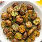 Vertical Pinterest pin showing red and Yukon Gold roasted baby potatoes piled in a large white ceramic bowl and garnished with chopped parsley and fresh herbs. Fresh parsley, rosemary, a black-and-white checkered towel, and a glass container of golden olive oil surround the bowl on a light surface. A white text box at the top reads "Garlic Herb Oven-Roasted Potatoes" in black lettering with a thin gold line between the two lines of text. "THEFORKEDSPOON.COM" appears along the bottom.
