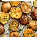 Vertical Pinterest pin featuring a close-up of golden roasted red and Yukon Gold baby potatoes on a silver baking sheet, topped with chopped green herbs, black pepper, and visible pieces of roasted garlic. A white text box across the upper portion reads "Garlic Herb Oven-Roasted Potatoes" in black lettering with a thin gold line between the two lines of text. "THEFORKEDSPOON.COM" appears along the bottom.