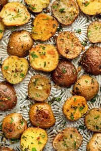 Close overhead view of roasted red and Yukon Gold baby potatoes scattered across a silver patterned baking sheet, with golden cut sides, browned skins, chopped parsley, rosemary, thyme, black pepper, and visible pieces of roasted garlic.