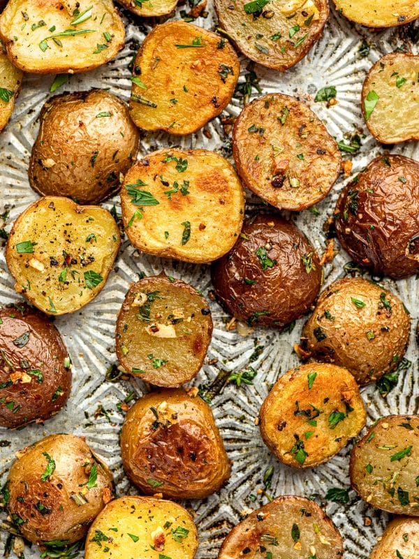 Close overhead view of roasted red and Yukon Gold baby potatoes scattered across a silver patterned baking sheet, with golden cut sides, browned skins, chopped parsley, rosemary, thyme, black pepper, and visible pieces of roasted garlic.