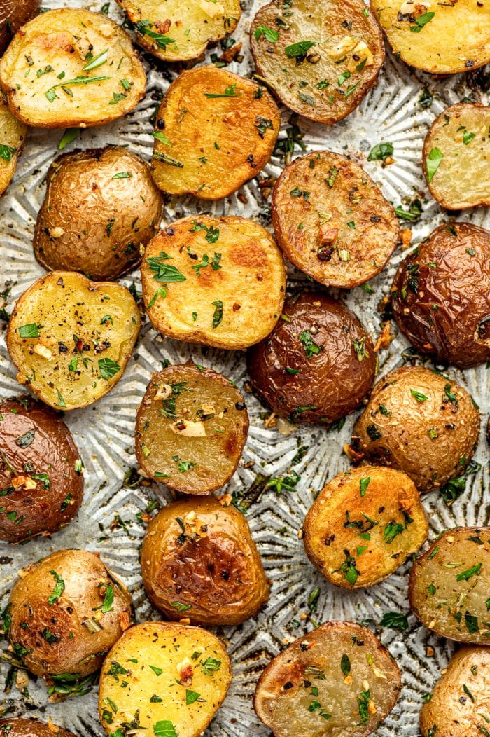 Close overhead view of roasted red and Yukon Gold baby potatoes scattered across a silver patterned baking sheet, with golden cut sides, browned skins, chopped parsley, rosemary, thyme, black pepper, and visible pieces of roasted garlic.