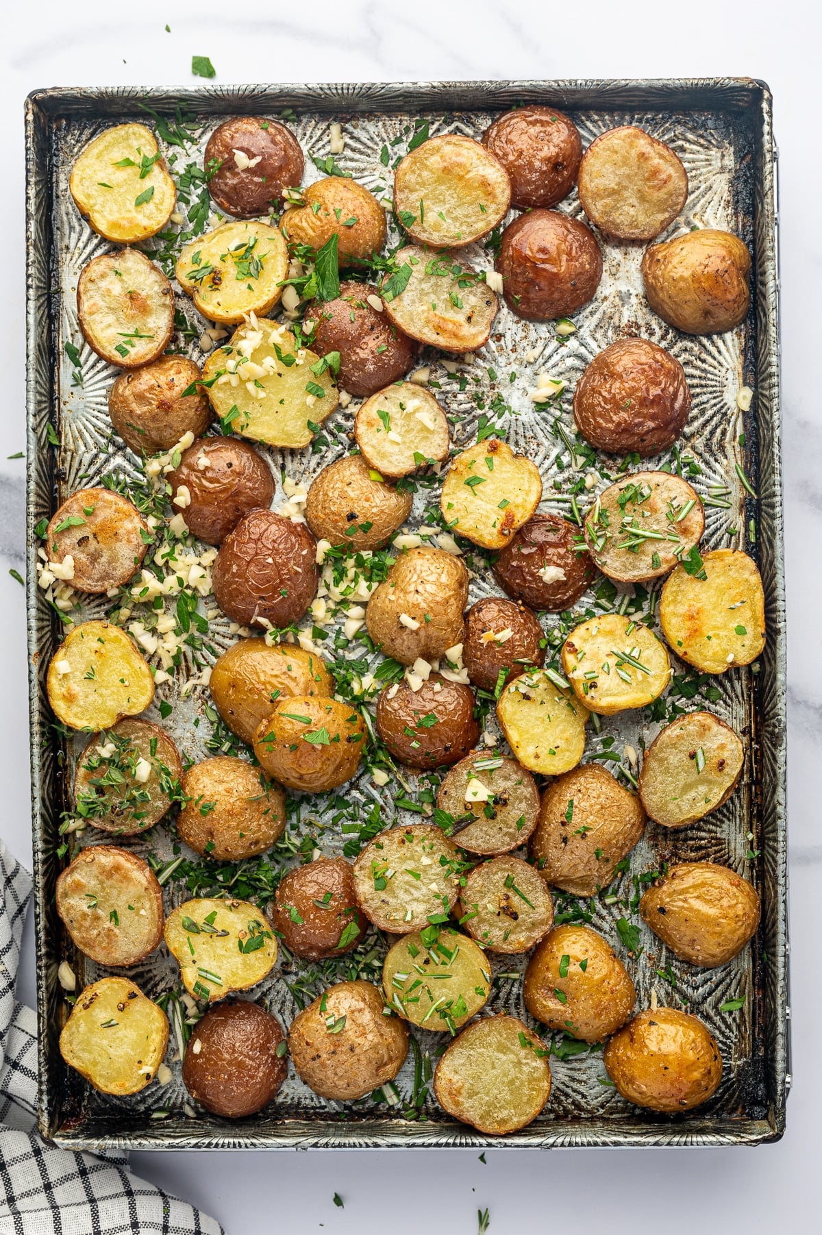 Overhead view of partially roasted red and Yukon Gold baby potatoes on a rectangular silver metal baking sheet, scattered with fresh minced garlic, chopped parsley, rosemary, and thyme. Some potato cut sides are lightly golden, and a black-and-white checkered towel is visible at the lower left.
