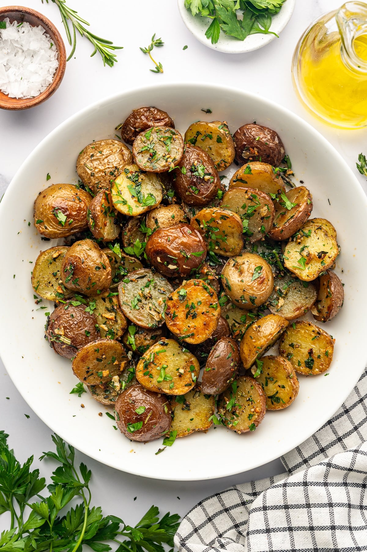 Overhead view of garlic roasted red and Yukon Gold baby potatoes in a large white ceramic bowl, garnished with chopped parsley, rosemary, and thyme. The bowl is surrounded by a small brown bowl of flaky salt, fresh rosemary sprigs, a small white bowl of parsley, a clear glass cruet filled with golden olive oil, loose parsley, and a black-and-white checkered kitchen towel on a white surface.