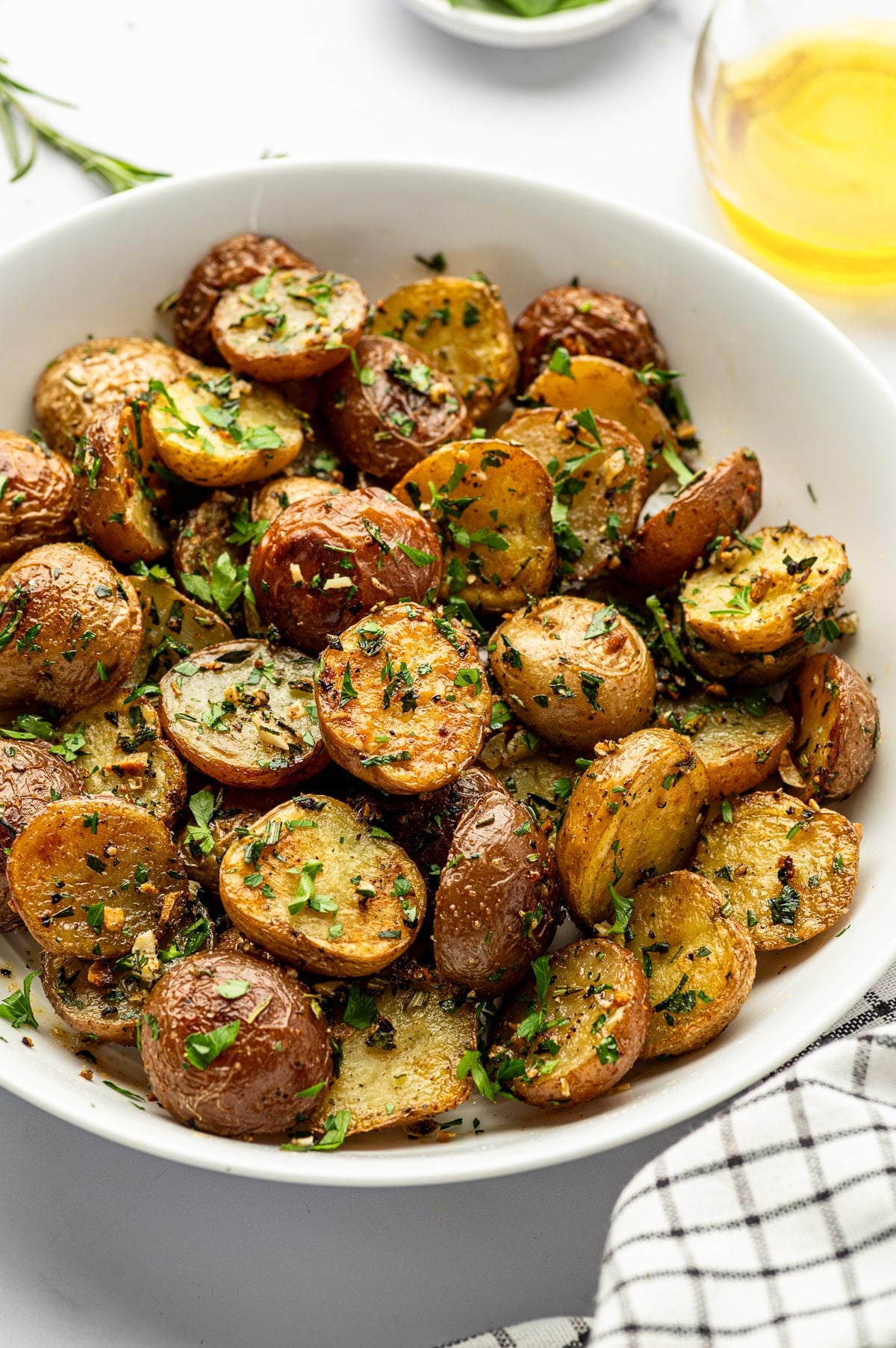 Close angled view of red and Yukon Gold garlic roasted baby potatoes piled in a large white ceramic serving bowl, with golden cut surfaces and browned skins topped with chopped parsley, rosemary, thyme, and pieces of roasted garlic. A clear glass container of golden olive oil is visible in the upper right, fresh rosemary appears at the left, and a black-and-white checkered towel is draped along the lower right.
