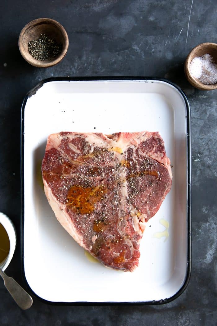 Raw T-Bone steak on a white tray seasoned with salt and pepper.