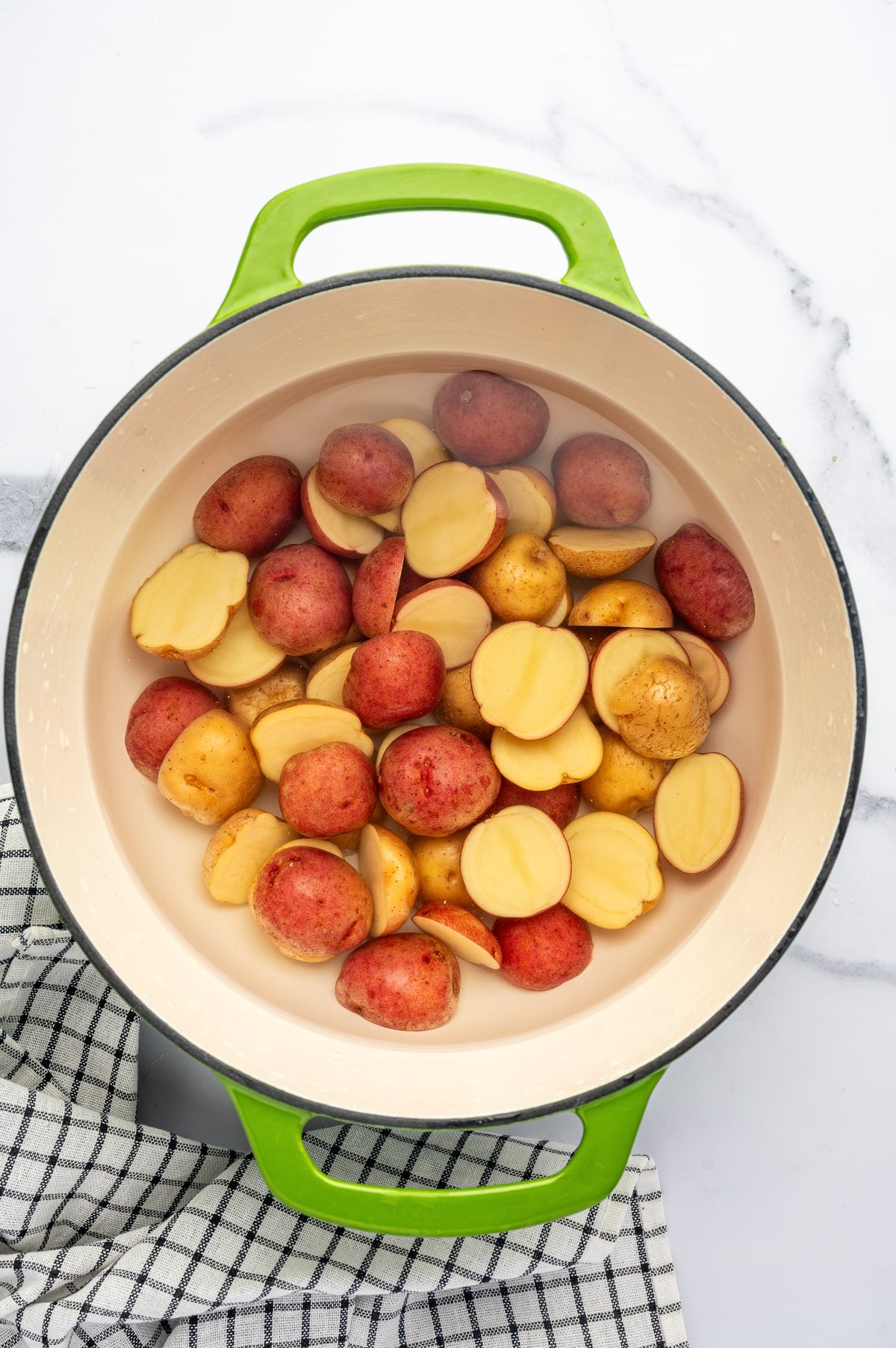 Overhead view of halved red and Yukon Gold baby potatoes submerged in water inside a bright green enameled Dutch oven with a cream-colored interior, set on a white marble surface with a black-and-white checkered kitchen towel underneath.