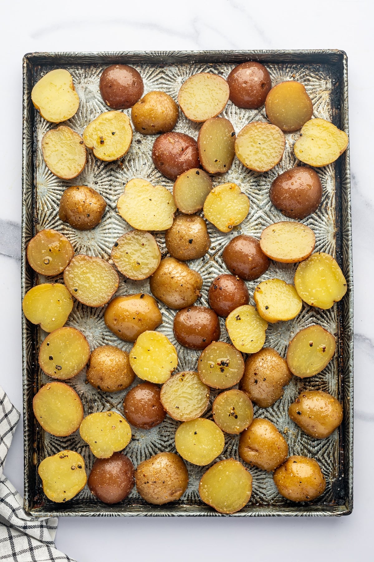 Overhead view of halved red and Yukon Gold baby potatoes arranged in a single layer on a rectangular silver metal baking sheet with dark edges, lightly coated with olive oil, garlic powder, salt, and black pepper. A black-and-white checkered towel is partially visible at the lower left.