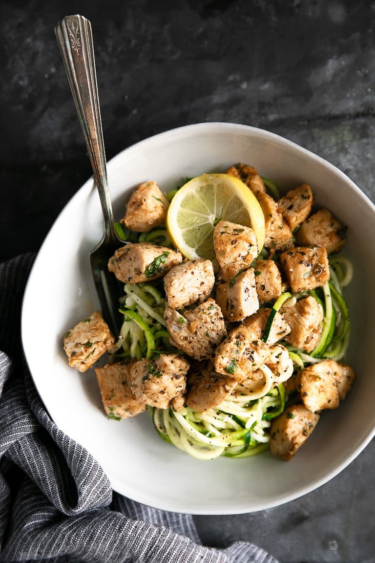 Overhead image of a large white serving bowl with zucchini noodles and garlic butter chicken chunks seasoning with pepper and minced parsley.
