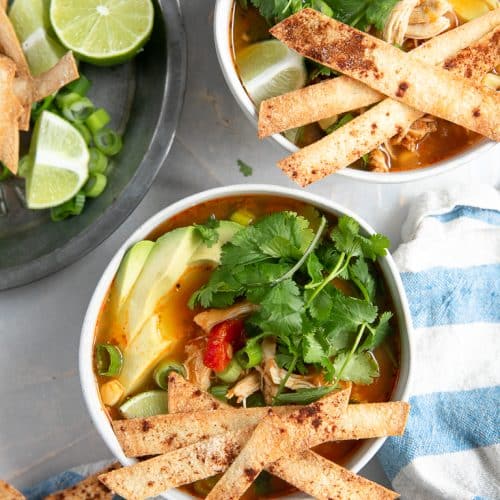 Overhead image of two white bowls filled with chicken enchilada soup and garnished with homemade baked tortilla strips, cilantro, and avocado.
