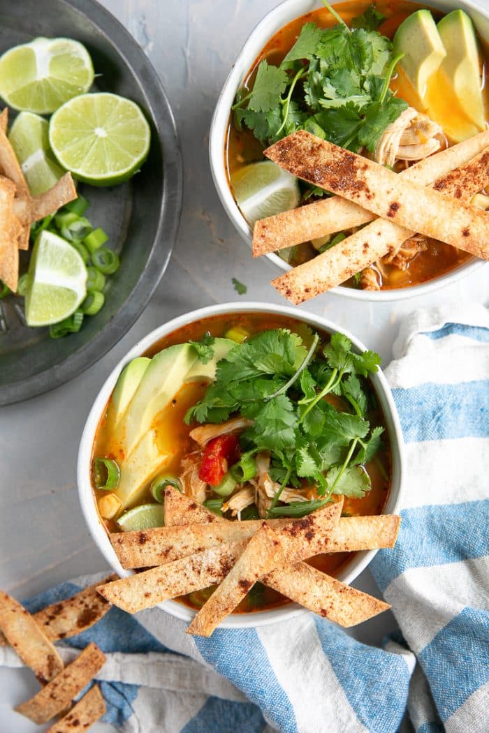 Overhead image of two white bowls filled with chicken enchilada soup and garnished with homemade baked tortilla strips, cilantro, and avocado.