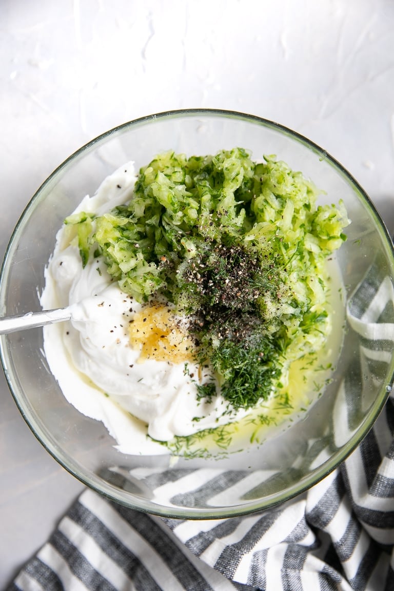 Glass bowl filled with plain Greek yogurt, lemon juice, cucumber, garlic, fresh dill, salt, and pepper.