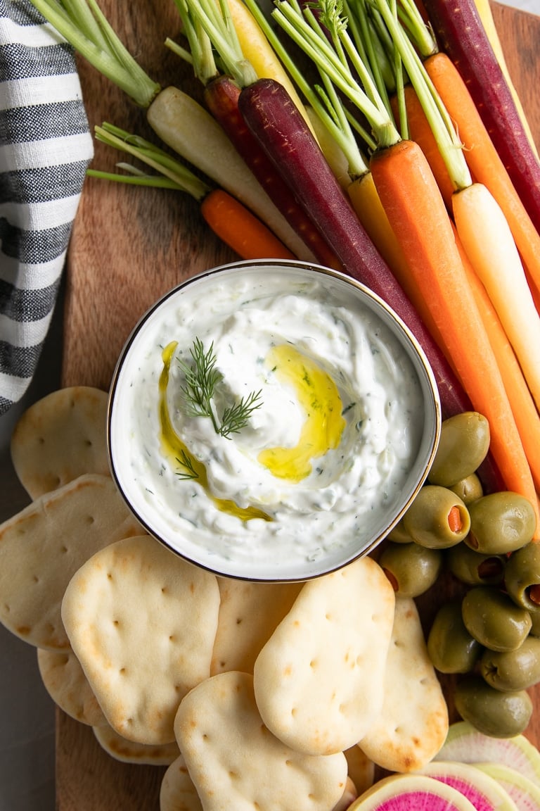 Close-up image of authentic tzatziki sauce drizzled with olive oil and surrounded by rainbow carrots and mini pita.