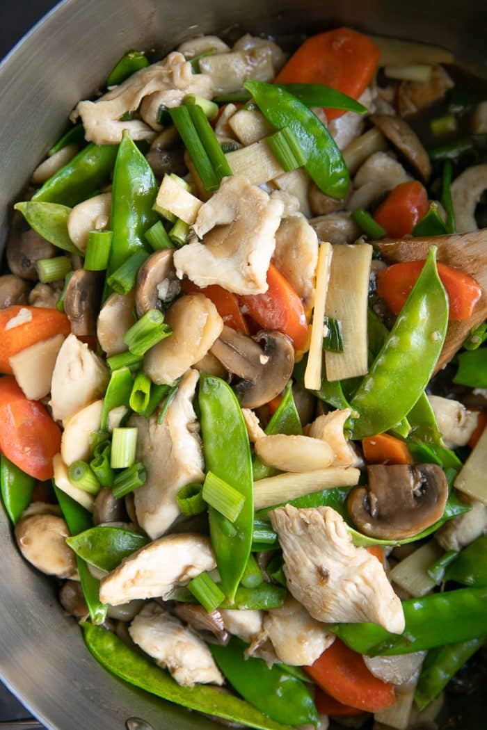 Large pan filled with stir-fried chicken, mushrooms, snow peas, carrots, water chestnuts, green onion, and chicken.
