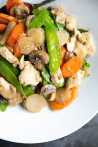 Moo Goo Gai Pan made with snow peas, water chestnuts, carrots, chicken, mushrooms, in a light sauce in a white shallow bowl.