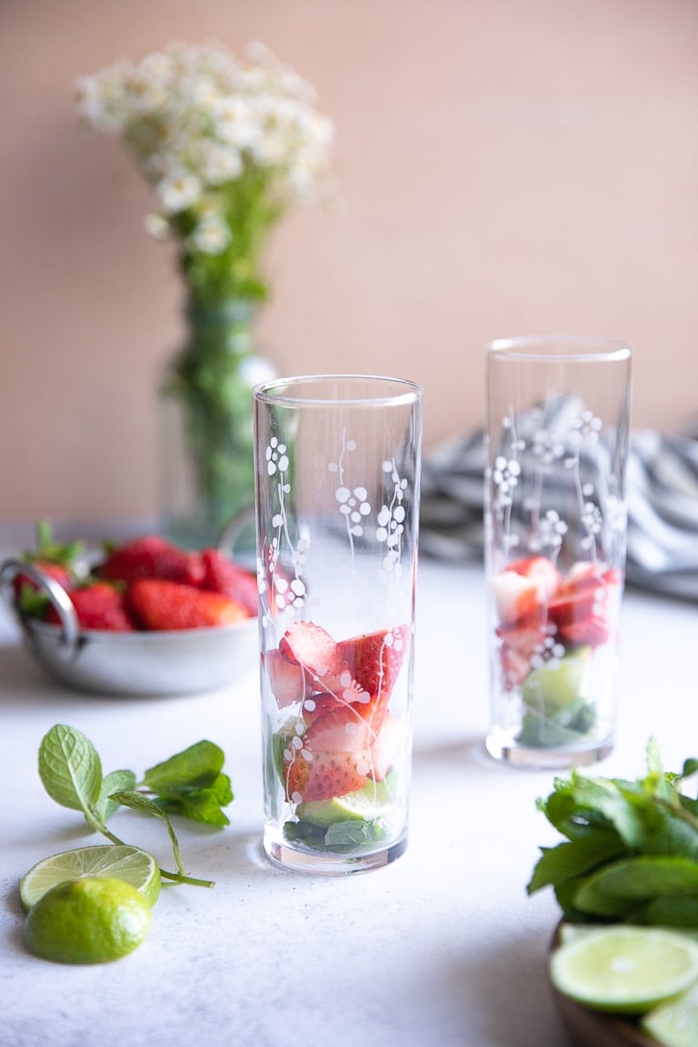 Highball glasses filled with mint, lime wedges, and chopped strawberries.