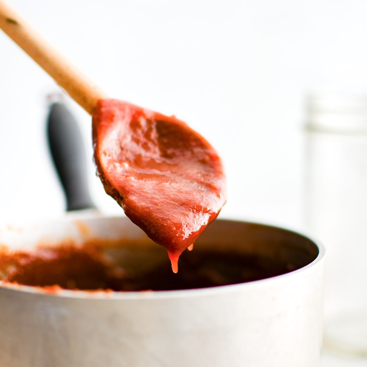 Homemade BBQ sauce dripping off a large wooden spoon back into a medium saucepot.