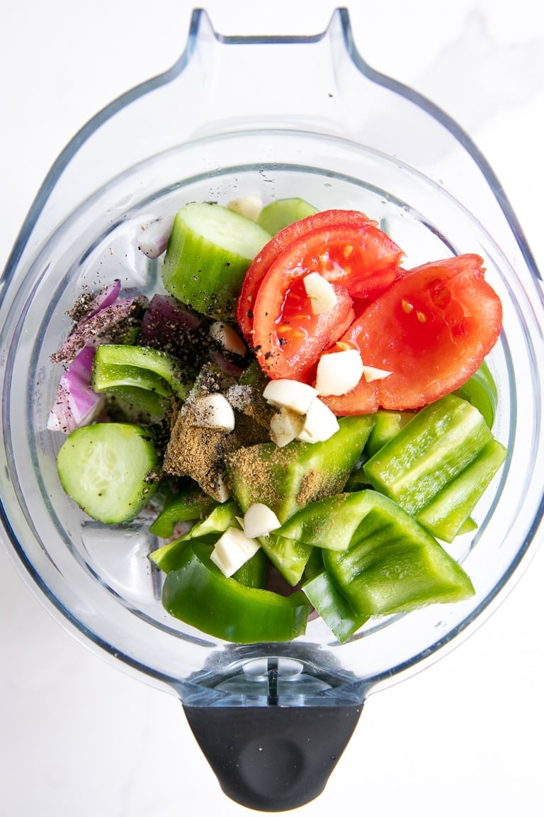 Blender filled with chopped war vegetables like tomatoes, cucumber, bell pepper, red onion.