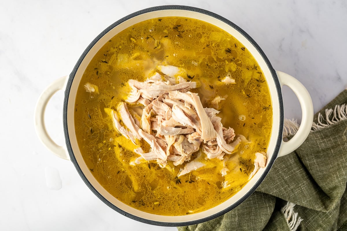 Shredded chicken piled in the center of a Dutch oven filled with golden chicken broth, tender carrots, celery, onions, and herbs.