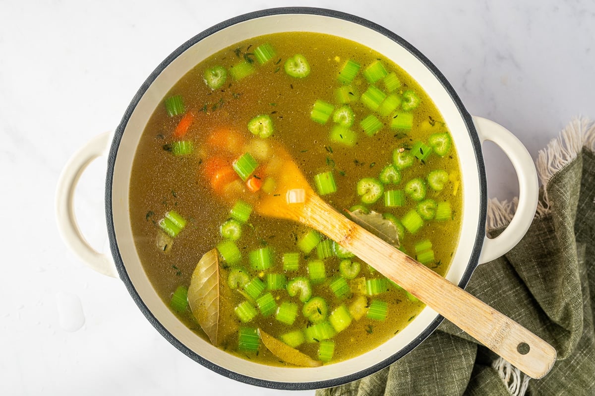 Golden homemade chicken broth added to carrots, celery, and onions in a white Dutch oven with bay leaves and thyme visible in the soup.