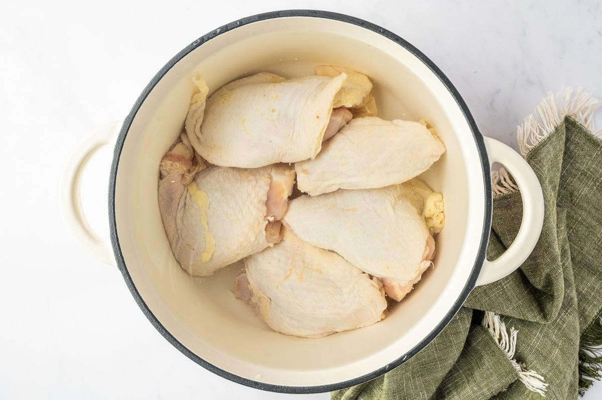 Bone-in, skin-on chicken thighs and boneless chicken breasts submerged in water in a large white Dutch oven to begin making homemade chicken broth for chicken noodle soup.
