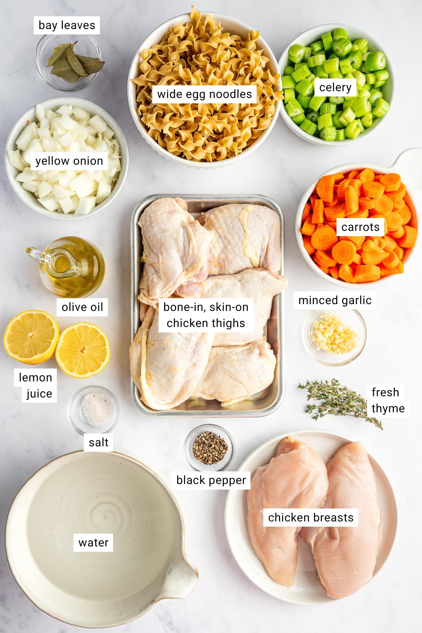 Ingredients for homemade chicken noodle soup arranged and labeled, including bone-in skin-on chicken thighs, chicken breasts, wide egg noodles, carrots, celery, yellow onion, minced garlic, fresh thyme, bay leaves, lemon, olive oil, salt, black pepper, and water.
