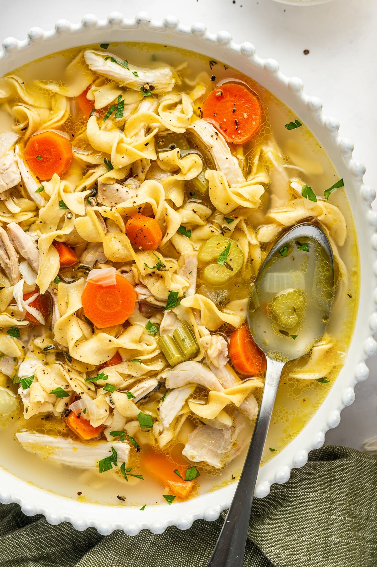 Bowl of chicken noodle soup with a metal soup spoon resting in the golden broth among wide egg noodles, shredded chicken, sliced carrots, celery, onions, and parsley.